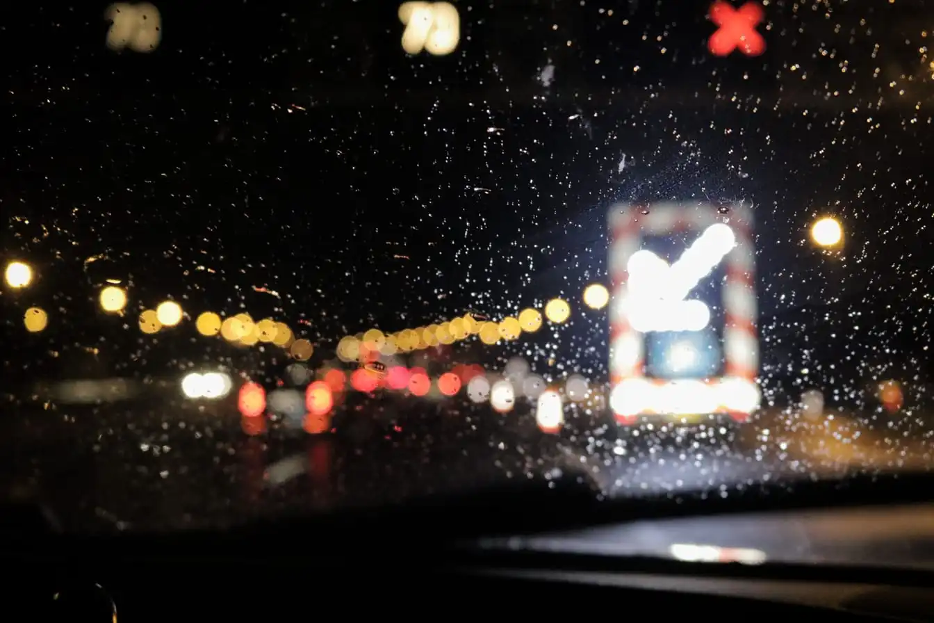 Blurry night traffic seen through a rain-soaked windshield, with glowing lights and reflections.