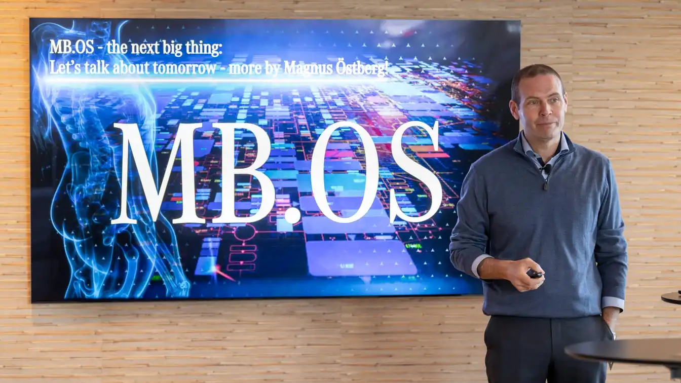 Magnus Östberg stands beside a presentation screen reading MB.OS, holding a presenter remote.