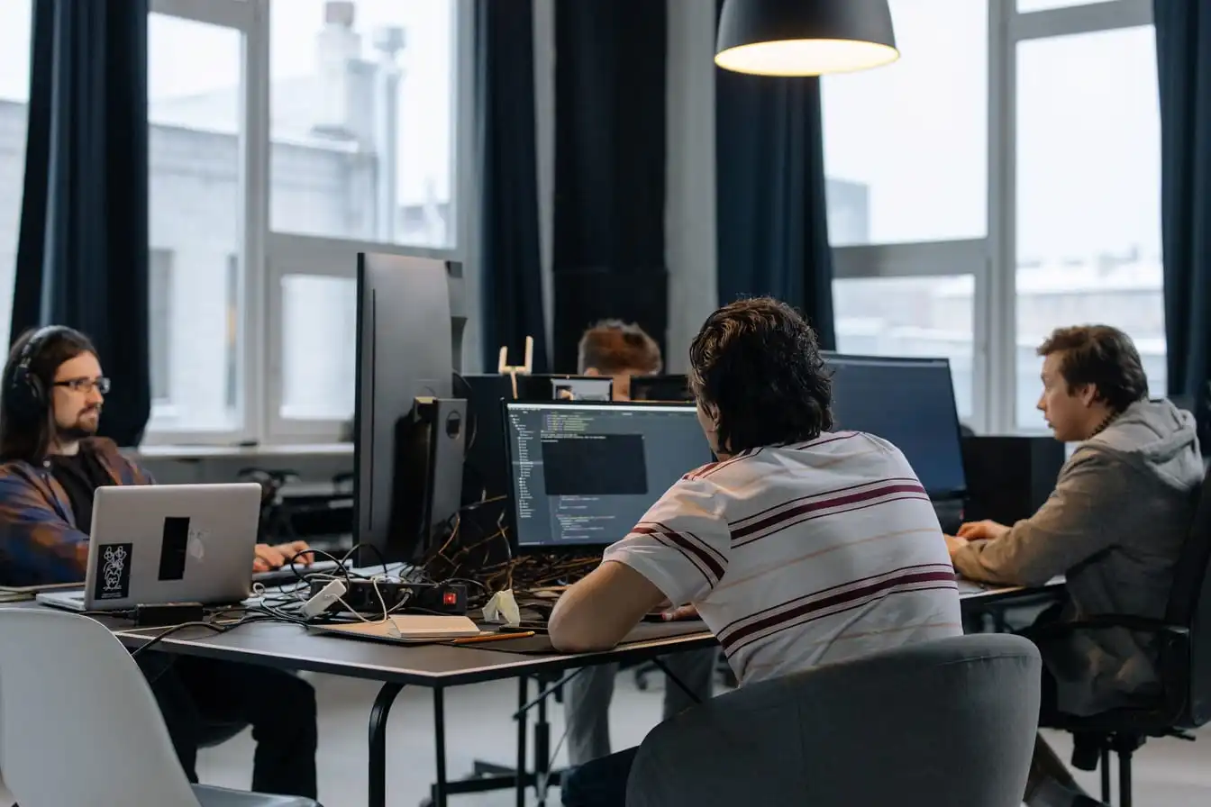 Four people at a shared desk in a bright open-plan office, working at laptops and monitors, one screen showing lines of code.