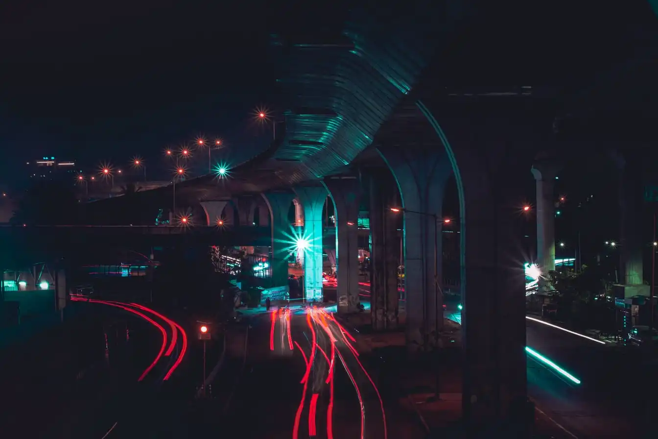 Long-exposure traffic light trails on a road passing beneath an elevated highway at night.