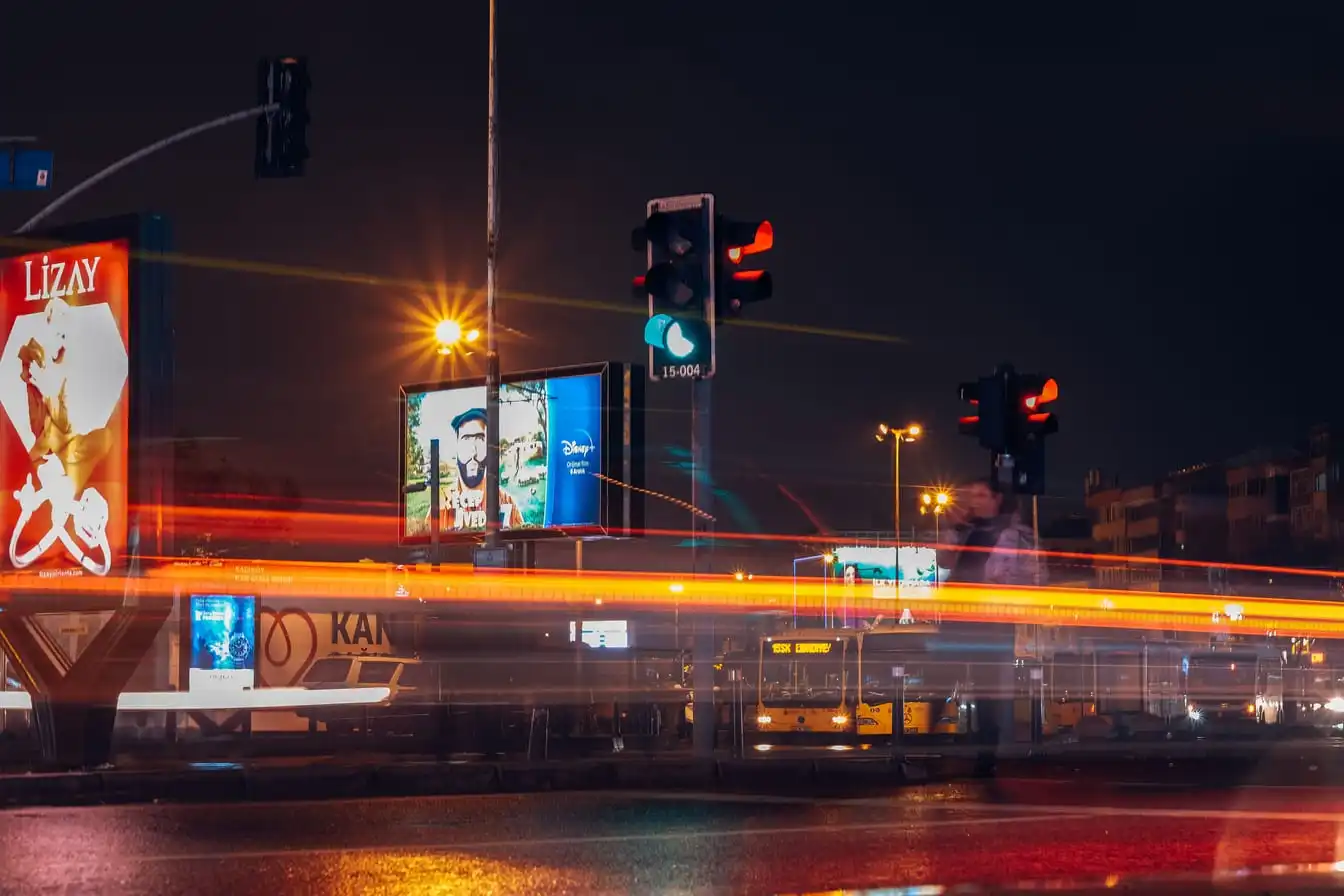 A city street at night in long exposure, with red and white light trails from passing traffic and a row of traffic signals overhead.