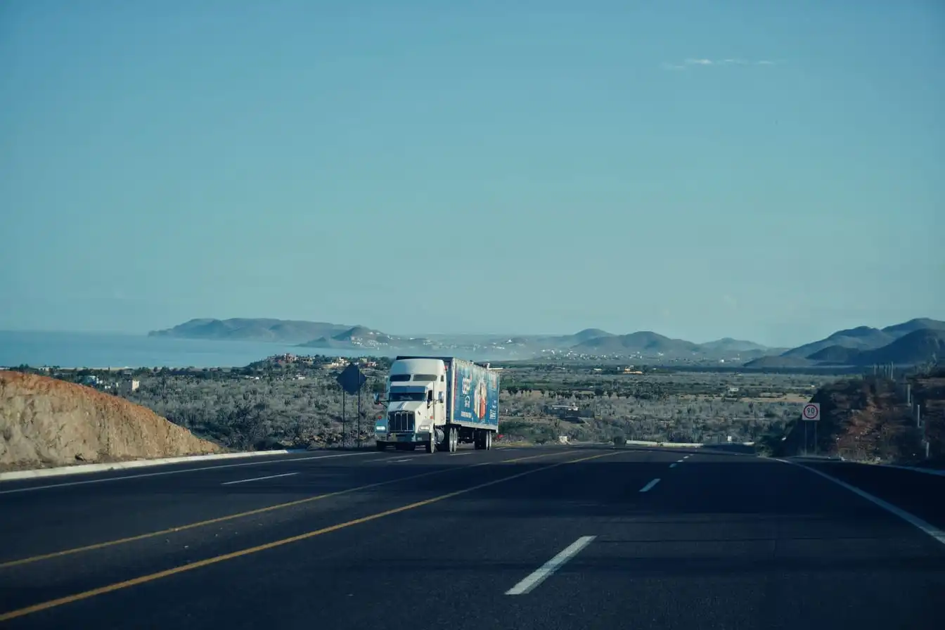 A semi-truck travels on a scenic highway with mountains in the background under a clear sky.